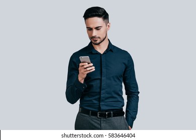 Typing quick message. Serious young man in shirt typing text message using his smart phone while standing against grey background