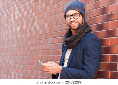 Typing text message. Side view of handsome young man in smart casual wear holding mobile phone while leaning at the brick wall - Shutterstock ID 224822299