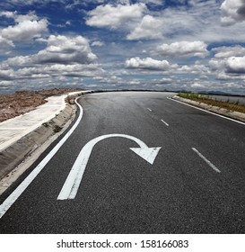 A U turn arrow traffic symbol imprint on a lonely asphalt road against a blue cloudy sky. 