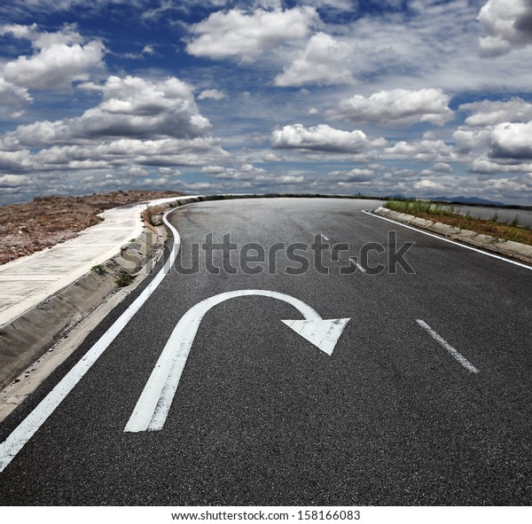 A U turn arrow traffic symbol\
imprint on a lonely asphalt road against a blue cloudy sky.\
