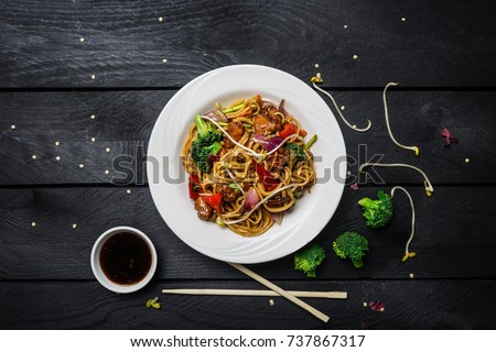 Udon stir fry noodles with chicken and vegetables in a white plate on black wooden background. With chopsticks and sauce
