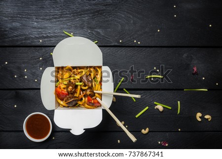 Udon stir fry noodles with meat and vegetables in a box on black background. With chopsticks and sauce.