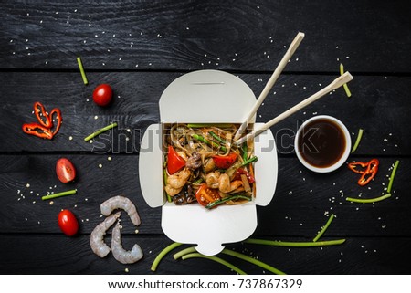 Udon stir fry noodles with seafood in a box on black background. With chopsticks and sauce.
