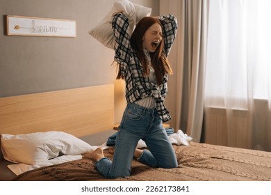 Ufa, Russia - 26 February 2021. Portrait of aggressive ginger young woman shouting and yelling with aggressive expression, throwing pillow sitting on bed in bedroom. Furious crazy redhead female - Shutterstock ID 2262173841