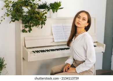 UFA, RUSSIA - 6 AUGUST 2021. Portrait of confident young woman pianist looking at camera sitting at classical white piano in classroom. Talented female musician performer preparing for classes 