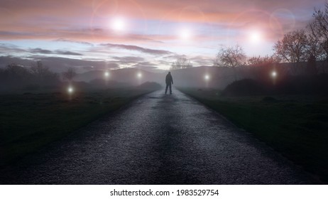 A UFO concept of glowing orbs, floating above a misty winters road just after sunset. With a silhouetted figure , back to camera, looking at the lights.     
