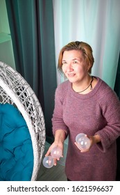 Ugly Aged Woman In A Sweater Juggling With White Balls Near White Wicker Hanging Chair In The Room With Curtains. Old Actress During Photoshoot In Studio