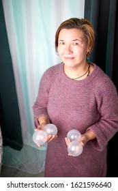 Ugly Aged Woman In A Sweater Juggling With White Balls Near White Wicker Hanging Chair In The Room With Curtains. Old Actress During Photoshoot In Studio