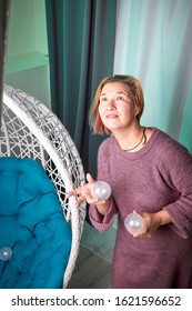 Ugly Aged Woman In A Sweater Juggling With White Balls Near White Wicker Hanging Chair In The Room With Curtains. Old Actress During Photoshoot In Studio