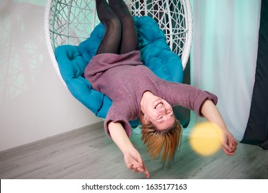 Ugly Aged Woman In A Sweater In A White Wicker Hanging Chair In The Room With Curtains. Old Model During Photoshoot In Studio