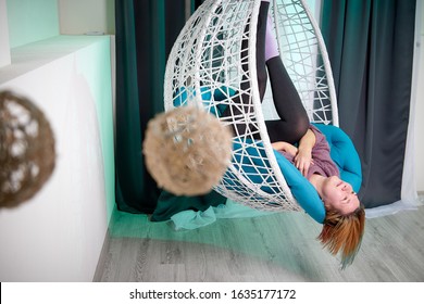 Ugly Aged Woman In A Sweater In A White Wicker Hanging Chair In The Room With Curtains. Old Model During Photoshoot In Studio