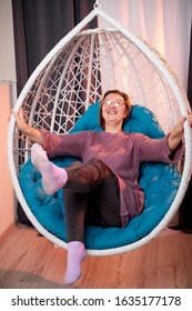 Ugly Aged Woman In A Sweater In A White Wicker Hanging Chair In The Room With Curtains. Old Model During Photoshoot In Studio