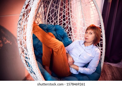 Ugly Girl In A White Shirt And Red Jeans On A White Wicker Hanging Chair In The Room With Curtains. Model During Photoshoot In Studio