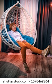 Ugly Girl In A White Shirt And Red Jeans On A White Wicker Hanging Chair In The Room With Curtains. Model During Photoshoot In Studio