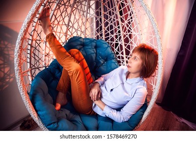 Ugly Girl In A White Shirt And Red Jeans On A White Wicker Hanging Chair In The Room With Curtains. Model During Photoshoot In Studio