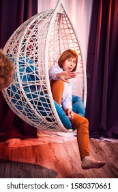 Ugly Girl In A White Shirt And Red Jeans On A White Wicker Hanging Chair In The Room With Curtains. Model During Photoshoot In Studio