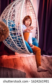 Ugly Girl In A White Shirt And Red Jeans On A White Wicker Hanging Chair In The Room With Curtains. Model During Photoshoot In Studio