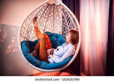 Ugly Girl In A White Shirt And Red Jeans On A White Wicker Hanging Chair In The Room With Curtains. Model During Photoshoot In Studio