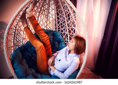 Ugly Girl In A White Shirt And Red Jeans On A White Wicker Hanging Chair In The Room With Curtains. Model During Photoshoot In Studio