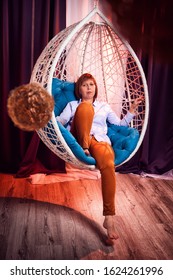 Ugly Girl In A White Shirt And Red Jeans On A White Wicker Hanging Chair In The Room With Curtains. Model During Photoshoot In Studio