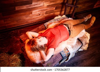 Ugly Pity Girl In Red Dress On The Couch With Hay In The Room Or In The Hayloft With Wooden Walls. Rustic Style In The Interior During Photoshoot