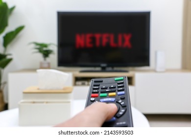 UK - April 5th 2022: First Person Perspective Of Hand Holding Remote Control Watching TV, Netflix Logo On Television Screen Out Of Focus, Depicting Home Life, Selective Focus