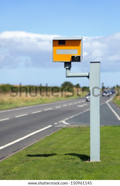UK static speed camera with speeding cars\
approaching on a rural road