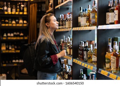 Ukraine. Kharkov Ocotber 2019 Young Beautiful Caucasian Girl In A Liquor Store Does Not Look At A Shelf With Bottles Of Whiskey. A Modern Woman Wearing Leather Jacket And Jeans Buying Alcohol