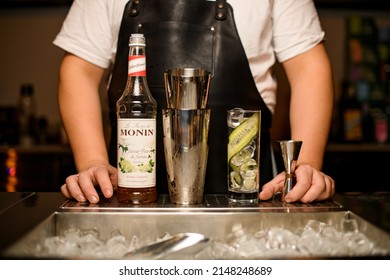 UKRAINE, KYIV - DECEMBER 5, 2021: Bottle of Monin elderflover syrup, steel shaker, jigger and high glass with ice and cucumber slices on the bar counter. Prepared to make cocktail. 