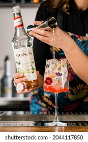 UKRAINE, KYIV - MARCH 12, 2021: Selective focus on beautiful glass with ice cubes in which the female hand pours liquid from steel jigger. Bottle of Monin brand in hand of barman