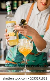 UKRAINE, KYIV - MARCH 12, 2021: beautiful glass with ice cubes stands on bar and woman gently bartender pours orange syrup from jigger into it and holds bottle of syrup by Monin brand in her hand