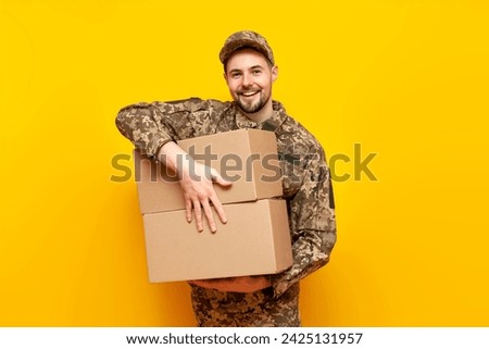 Ukrainian army soldier in pixel camouflage uniform holds a box on a yellow isolated background, Ukrainian military cadet carries and gives humanitarian aid and donation