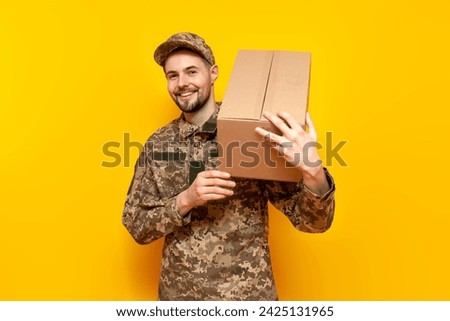 Ukrainian army soldier in pixel camouflage uniform holds a box on a yellow isolated background, Ukrainian military cadet carries and gives humanitarian aid and donation