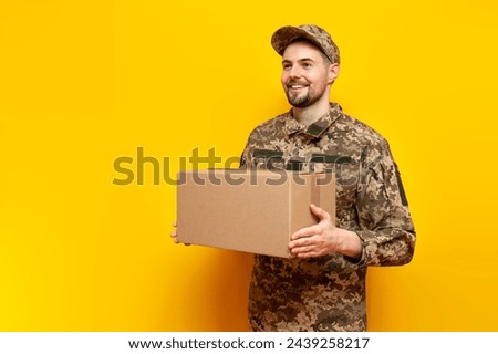 Ukrainian army soldier in pixel camouflage uniform holds a box on a yellow isolated background, Ukrainian military cadet carries and gives humanitarian aid