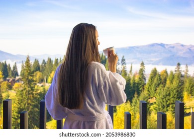 Ukrainian brunette girl with long hair from back in white bathrobe holding cup of tea, coffee drink standing on balcony, breathing in clean morning air. Mountains view. Spa recreation. Good morning. - Powered by Shutterstock