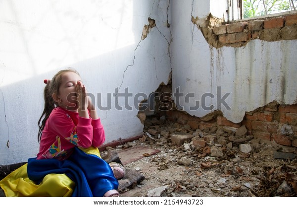 A\
Ukrainian child prays for an end to the war. Consequences of the\
Russian war against Ukraine. War in Ukraine. Children against war.\
Peace concept. A child in front of a war-torn\
house.