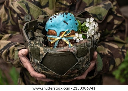 Ukrainian Easter Cake in A Hands of a Soldier in a Military Helmet. Army of Ukraine celebrate of Easter. War in Ukraine. Easter Bread with Ukraine Symbols and spring flowers. Focus on glaze