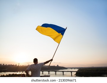 Ukrainian flag flutters in the wind against the sunset sky. A man standing on the roof of a house in Kyiv holds a large flag. National symbol of freedom and independence. Stop the war. Hope and Faith