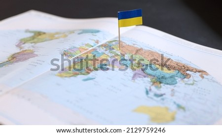 A Ukrainian flag stuck in the position of where Ukranine is located on a colored map of the world in an open atlas. Where is Ukraine? Location and situation in Eastern Europe.