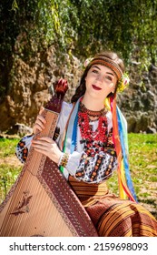 Ukrainian Musician In Authentic National Dress Sits By A Rock. Ukrainian Woman With A Bandura Musical Instrument Before Performing Folk Music