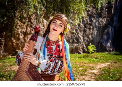 Ukrainian Musician In Authentic National Dress Sits By A Rock. Ukrainian Woman With A Bandura Musical Instrument Before Performing Folk Music