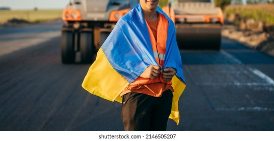 Ukrainian Smiling Young Worker With Ukrainian Flag On His Shoulders On The Background Of Road Works