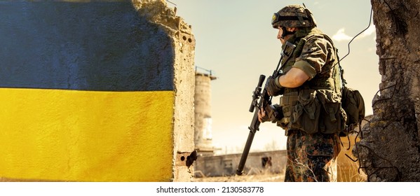 Ukrainian Soldier Military In The War With A Weapon In His Hands. The Flag Of Ukraine Is Painted On A Brick Wall. Relations Between Ukraine And Russia