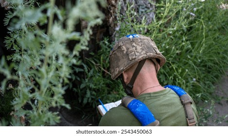 Ukrainian soldier seen from behind, crouched near a bunker entrance, wearing a helmet and green clothing, engaged in writing or noting something down, symbolizing the dedication amidst conflict - Powered by Shutterstock