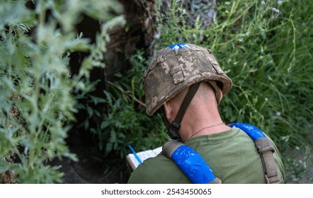 Ukrainian soldier seen from behind, crouched near a bunker entrance, wearing a helmet and green clothing, engaged in writing or noting something down, symbolizing the dedication amidst conflict - Powered by Shutterstock