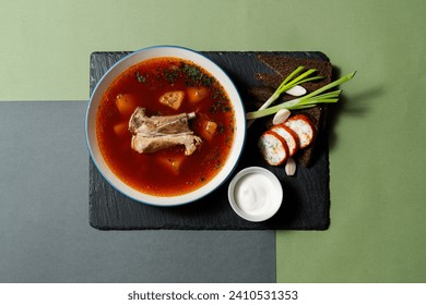 Ukrainian soup Borsch with beef on the bone in a deep plate. Toasts made of black bread, pieces of lard, green onions and a bowl with sour cream lie nearby. food lies on a slate board on a paper. - Powered by Shutterstock