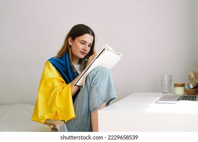 A Ukrainian teenage girl is wrapped in the Ukrainian flag   is drawing doing homework in the bedroom  A teenager studies at home  The concept studies during the war in Ukraine 