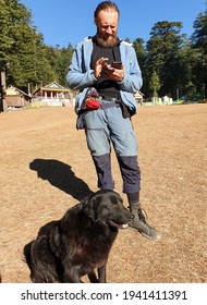 A Ukrainian Tourist Using His Phone In Outdoor And A Black Dog Sitting In Ground, Full Length Photo Of A Long Beard Man Standing In The Nature.