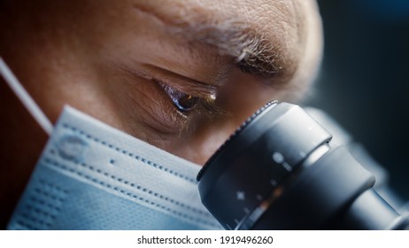 Ultra Macro Close Up Shot Of A Male Scientist Wearing Surgical Mask And Looking Into The Microscope. Microbiologist Working On Molecule Samples In Modern Laboratory With Technological Equipment.