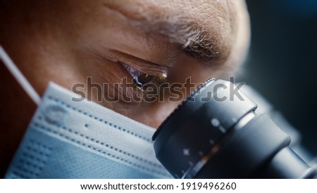 Ultra Macro Close Up Shot of a Male Scientist Wearing Surgical Mask and Looking into the Microscope. Microbiologist Working on Molecule Samples in Modern Laboratory with Technological Equipment.